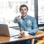 hey-you-portrait-happy-positive-bearded-young-freelancer-blue-jeans-shirt-are-sitting-cafe-working-laptop-with-toothy-smile-pointing-finger-you-looking-camera-indoor_416530-10708
